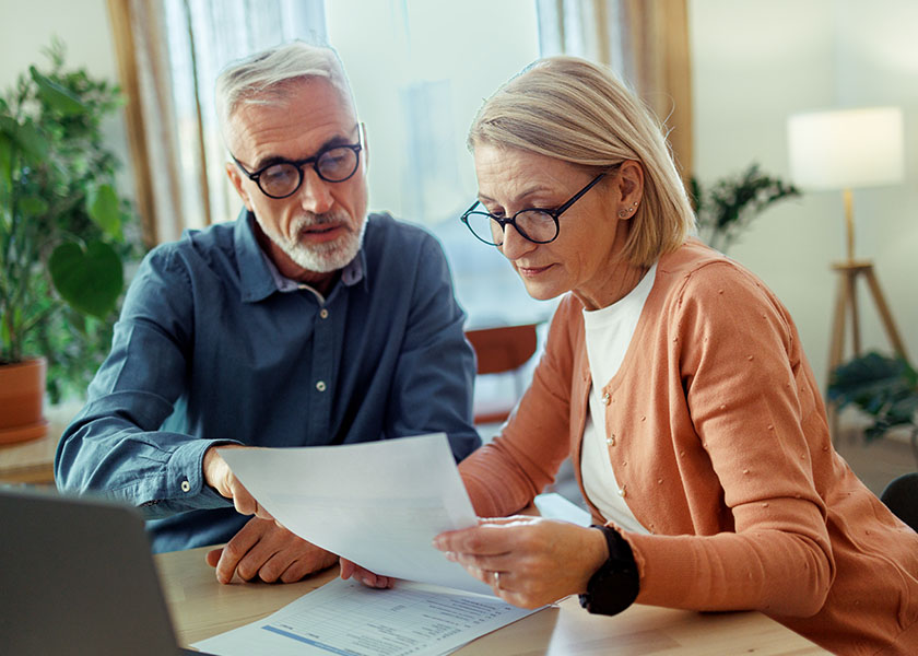 A mature couple sits at a table reviewing pages of information, a laptop nearby.